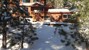 A two-story wooden cabin with a covered porch and a garage, surrounded by snow-covered trees.
