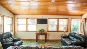 A room with wooden ceiling, large windows, a flat-screen TV, leather sofas, and a small wooden table with a plant.