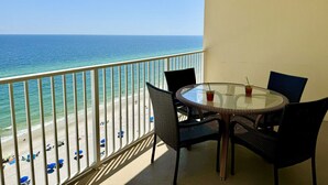 Een balkon met een ronde tafel en stoelen, met uitzicht op een strand met mensen en parasols.