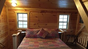 A cozy attic bedroom with a wooden sloped ceiling, two windows, a bed with a colorful quilt, and a small table. - Private Ravine View Luxury Amish-Built Cabin "The Frontier" (Genoa)