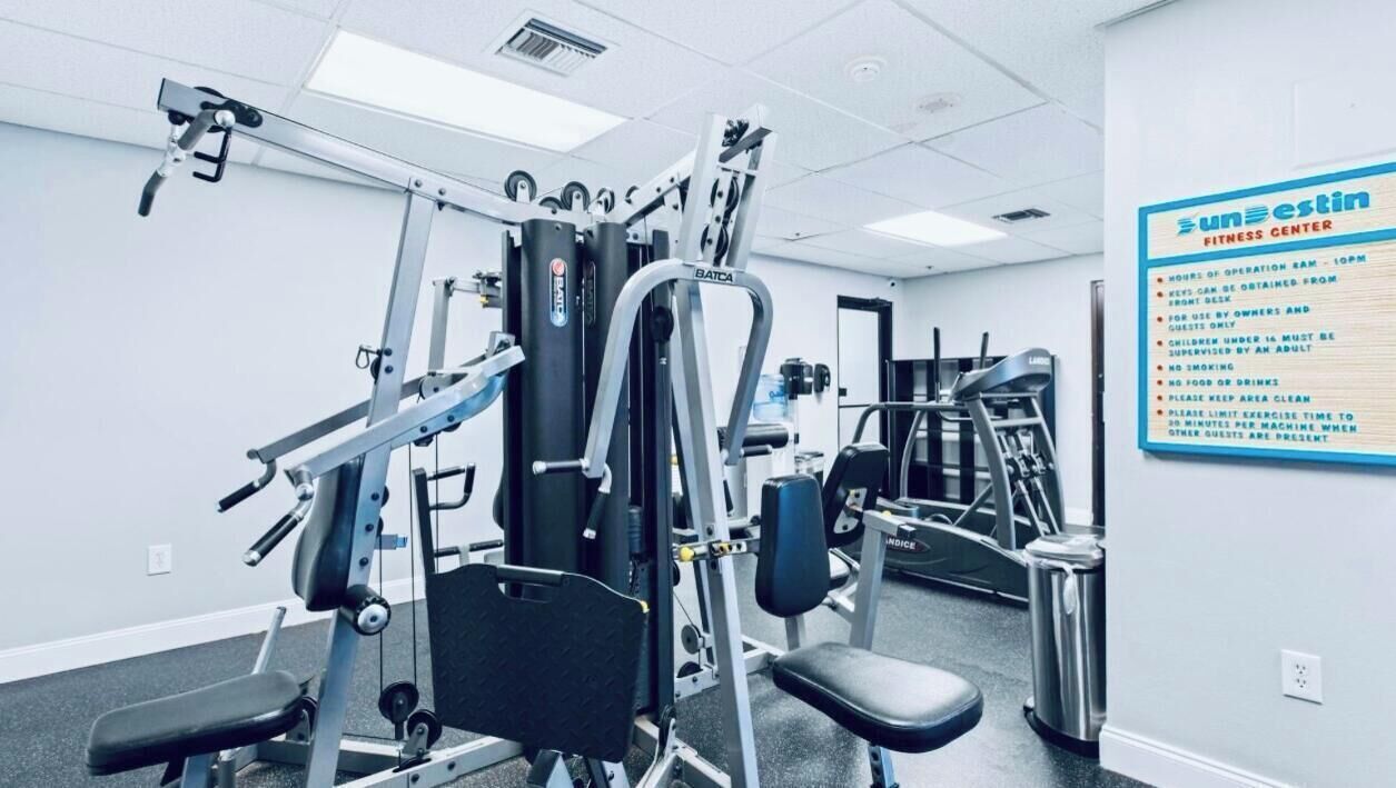 A fitness center with various exercise machines, a wall sign with operational hours, and a trash can.
