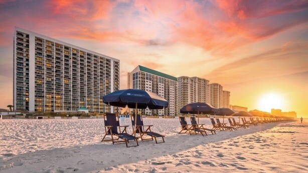 Una playa con filas de tumbonas y sombrillas, un edificio de hotel y una puesta de sol al fondo.