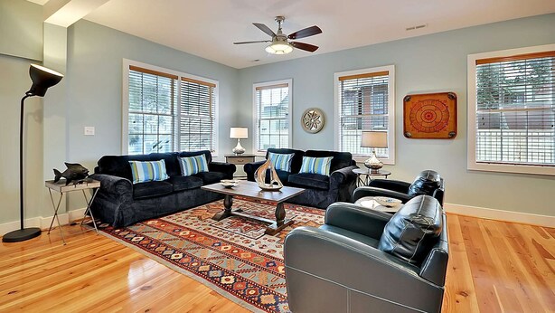 A living room with a dark-colored sofa set, a coffee table, and a patterned rug. There are two windows with blinds, a ceiling fan, and a decorative wall piece. - Airy & Bright Folly Condo; Close to Everything (Folly Beach)