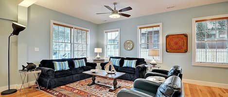 A living room with a dark-colored sofa set, a coffee table, and a patterned rug. There are two windows with blinds, a ceiling fan, and a decorative wall piece.