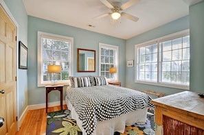 A bedroom with a bed, two bedside tables with lamps, a ceiling fan, and large windows with blinds.