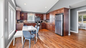 A modern kitchen with wooden cabinets, a stainless steel refrigerator, and a dining area with a white table and blue chairs. - Beachfront Chalet (Stratford)