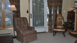 Una sala de estar con un sillón marrón, una silla de madera, una lámpara y una puerta con persianas.