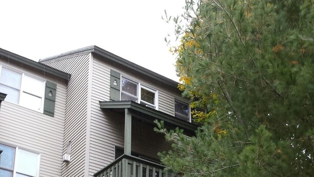 Un edificio de varios pisos con un balcón en el segundo nivel, una cerca de madera al frente y un árbol a la derecha.