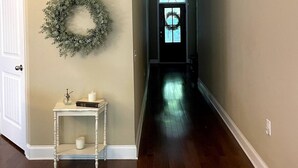 Un pasillo con suelo de madera, una puerta blanca, una pequeña consola blanca con una vela y un tarro de cristal, y una guirnalda en la pared.