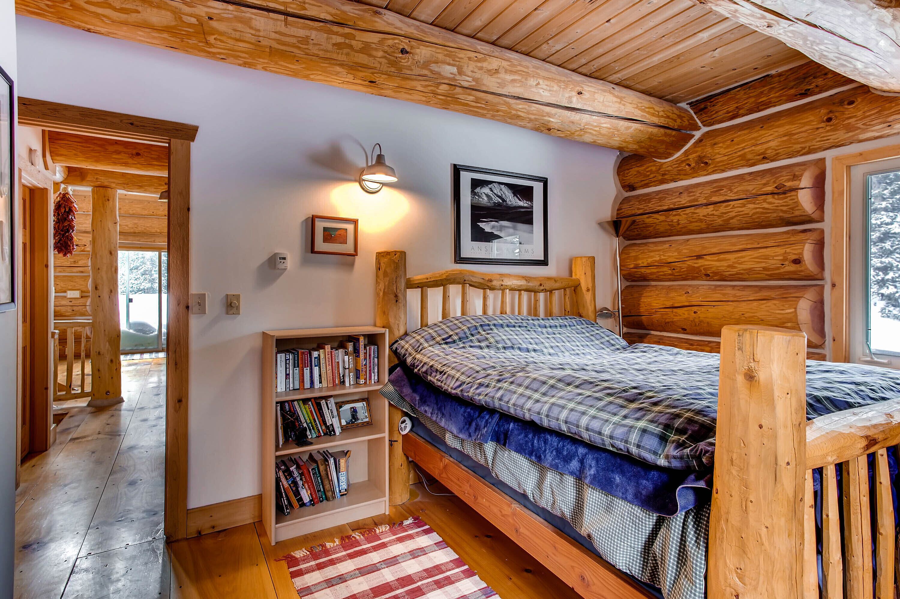 A cozy log cabin bedroom with a wooden bed, a bookshelf, a framed picture, and a window with a view of trees.