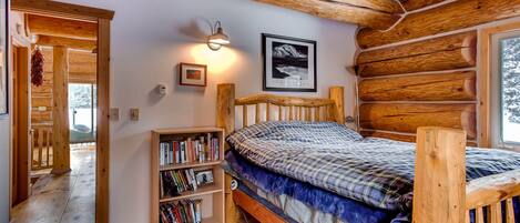 A cozy log cabin bedroom with a wooden bed, a bookshelf, a framed picture, and a window with a view of trees.