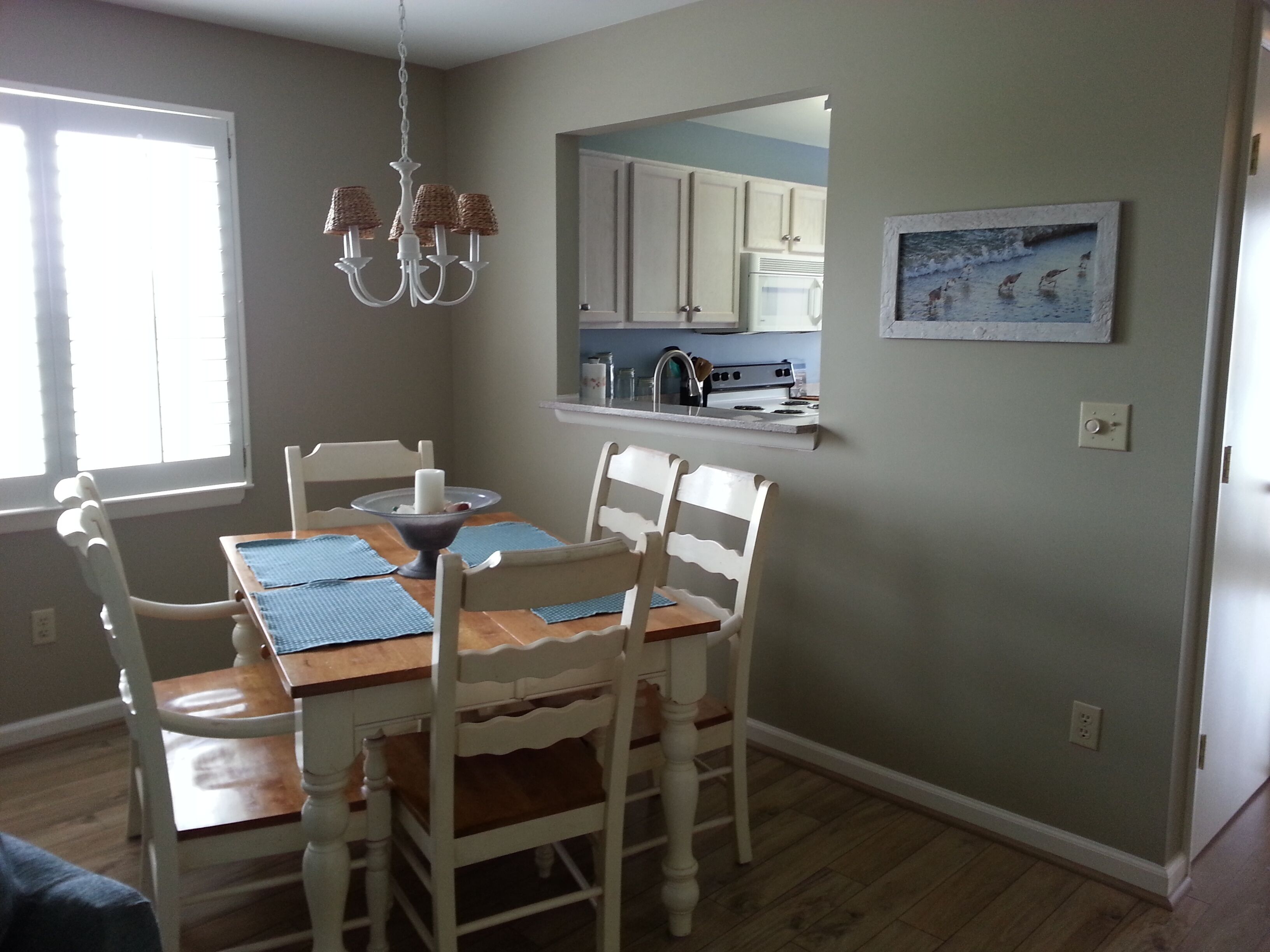 A dining area with a wooden table, white chairs, a chandelier, and a kitchen in the background.