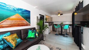 Un salon moderne avec un plancher en bois, un canapé en cuir noir agrémenté de coussins colorés, une table basse blanche et un grand tableau représentant une jetée en bois. On aperçoit, au fond, la cuisine avec ses armoires blanches et son comptoir noir.