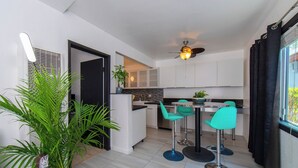 Une cuisine moderne avec des meubles blancs, un plan de travail noir et un ventilateur de plafond. On y trouve deux tabourets de bar turquoise et une table à manger.