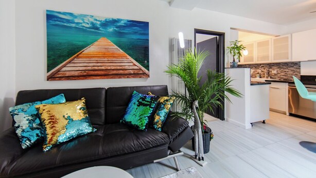 Un salon moderne avec un canapé en cuir noir, des coussins colorés, un grand tableau représentant un paysage marin et une cuisine en arrière-plan.