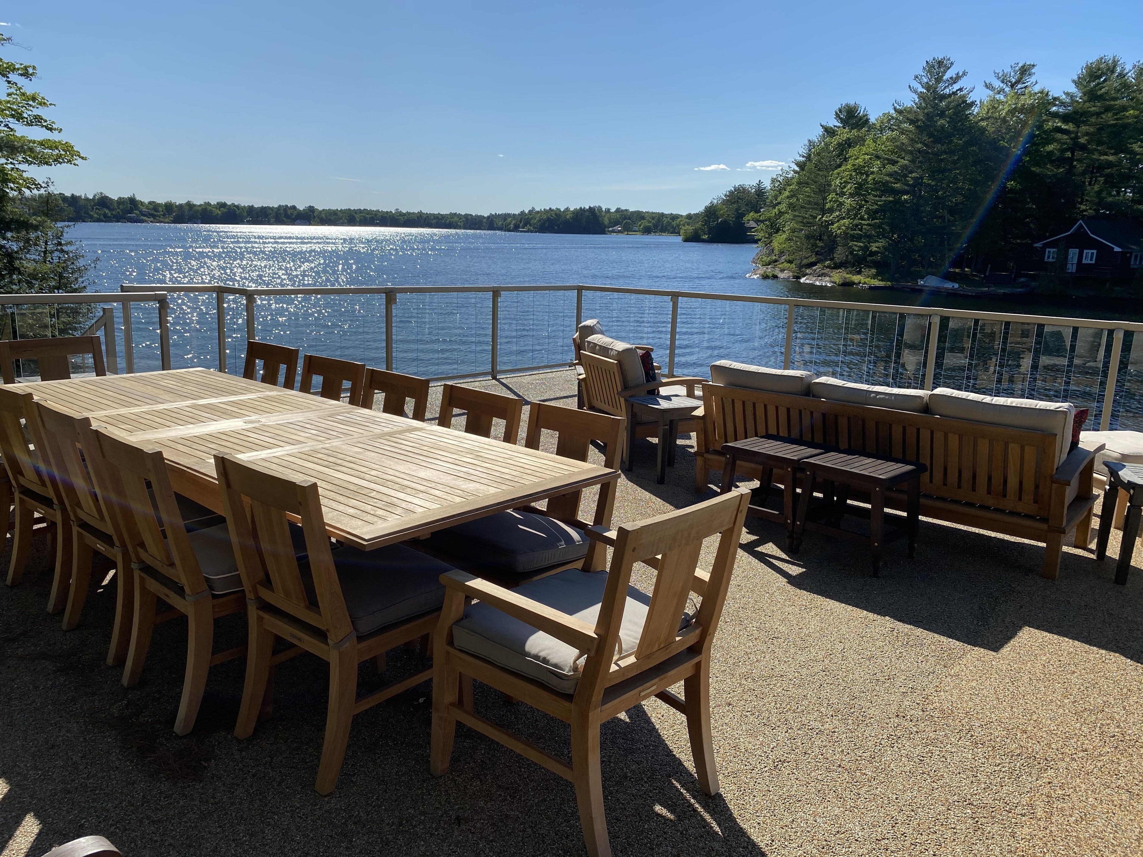 A wooden deck with a table and chairs overlooking a lake, surrounded by trees and a house in the background.