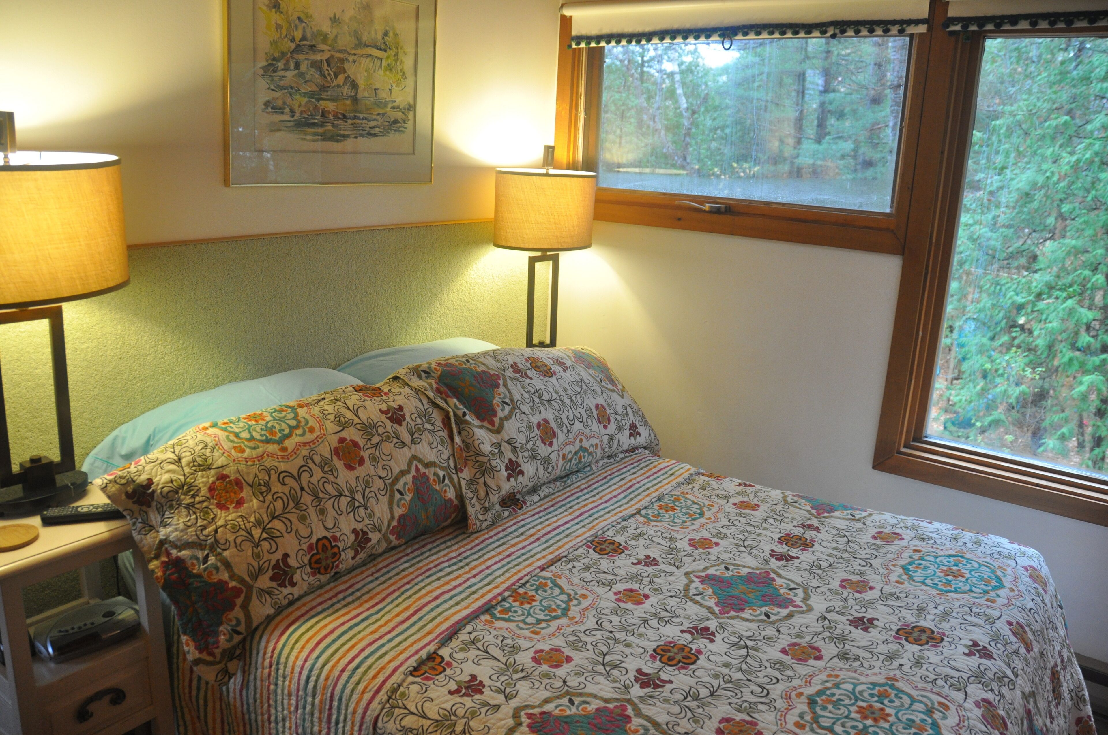 A bedroom with a bed, two bedside lamps, a window with a view of trees, and a painting on the wall.