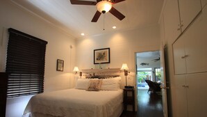 Un dormitorio con cama, lámparas de noche, ventilador de techo, un cuadro enmarcado y vistas a un balcón con mobiliario exterior.