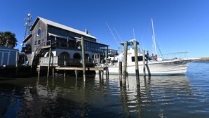 Un muelle de madera con un barco amarrado a un espigón, un edificio con balcón y un cielo azul despejado.