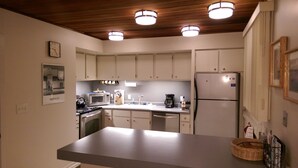 A kitchen with white cabinets, a microwave, a stove, a refrigerator, and a sink. There are three ceiling lights and a clock on the wall.
