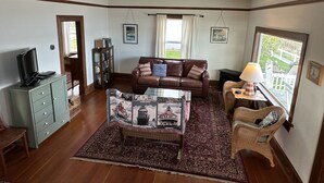 A living room with a brown leather sofa, a patterned coffee table, a wicker chair, a television on a cabinet, and a view of a deck through a window.