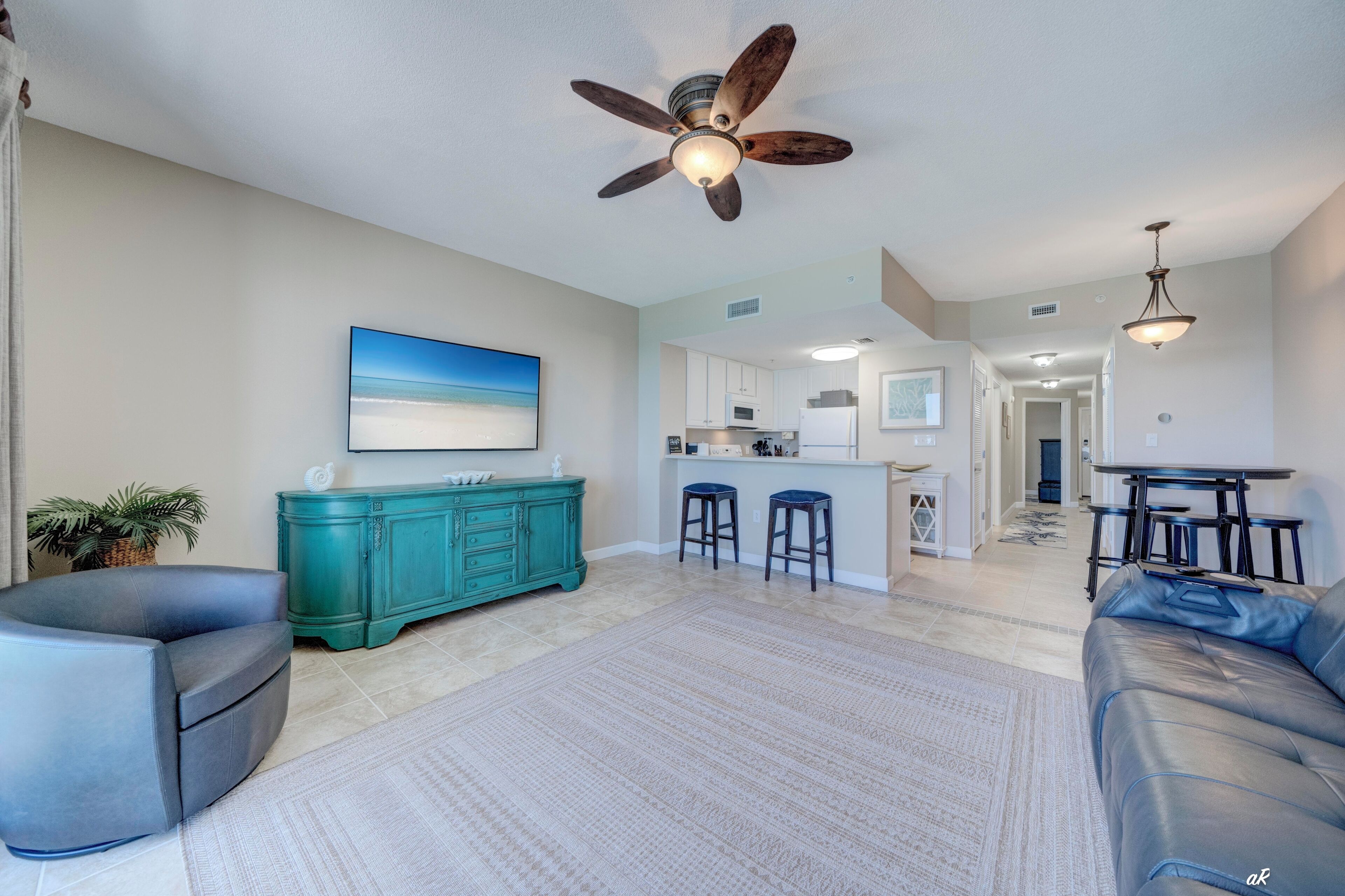 A spacious living area with a ceiling fan, a teal cabinet, a sofa, and a kitchen in the background.
