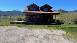 A rustic wooden cabin with a metal roof, situated in a grassy field with mountains in the background.