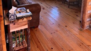 A log cabin interior with a fireplace, leather armchairs, a wooden coffee table, and large windows.