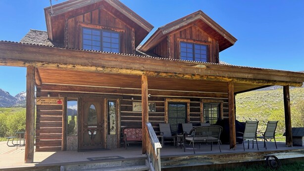 A rustic log cabin with a covered porch, outdoor seating, and a scenic mountain backdrop.