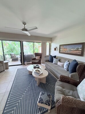 A modern living room with a sofa, coffee table, and armchairs, a ceiling fan, and a large window with a view of greenery.