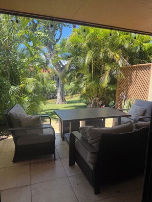 An outdoor patio with wicker furniture, a table, and a view of a lush garden with palm trees and a wooden fence.