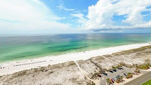 Luftaufnahme eines Strandes mit blauem Himmel, sandigem Ufer und einem Parkplatz mit Autos und einigen Personen.