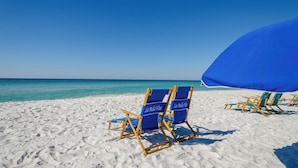 A beach with white sand and clear blue water. There are blue beach chairs and a blue umbrella.