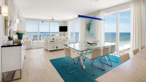 A modern hotel room with a glass dining table, white furniture, a large window with a view of the ocean, and a ceiling fan.