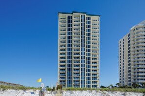 Un hôtel de grande hauteur doté de nombreux balcons, une plage de sable en premier plan et un ciel bleu dégagé.