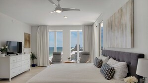 A modern hotel room with a large bed, a TV, a ceiling fan, and a view of the beach.