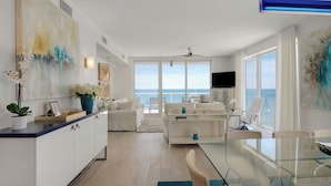 A modern living room with a large window overlooking the sea, a glass dining table, and a kitchen area with white cabinetry.