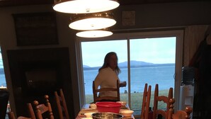 Taula de dinar amb un bol de pomes, cadires de fusta i vista al mar a través d'una finestra gran.