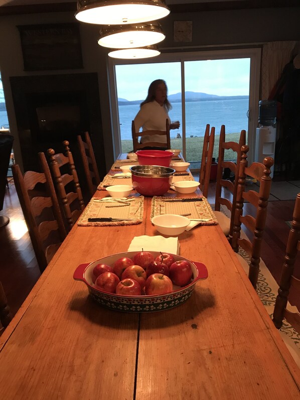 Une table à manger avec un saladier de pommes, des chaises en bois et une vue sur la mer par une grande fenêtre.