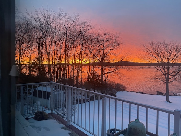 A balcony with a railing, a fire hydrant, and a snowy landscape with trees and a sunset over a body of water.