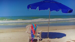 Una silla de playa con una toalla colorida y una sombrilla azul sobre una playa arenosa, con olas del océano al fondo.