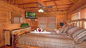 Een slaapkamer in een blokhut met een bed, houten hoofdbord, plafondventilator, televisie en houten jaloezieën.