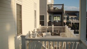 Une terrasse en bois avec des fauteuils Adirondack et une petite table, une balustrade blanche et une vue sur un immeuble de deux étages doté d’un balcon.