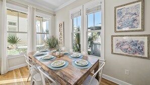 A dining area with a wooden table set for six, white chairs, and large windows with white curtains.