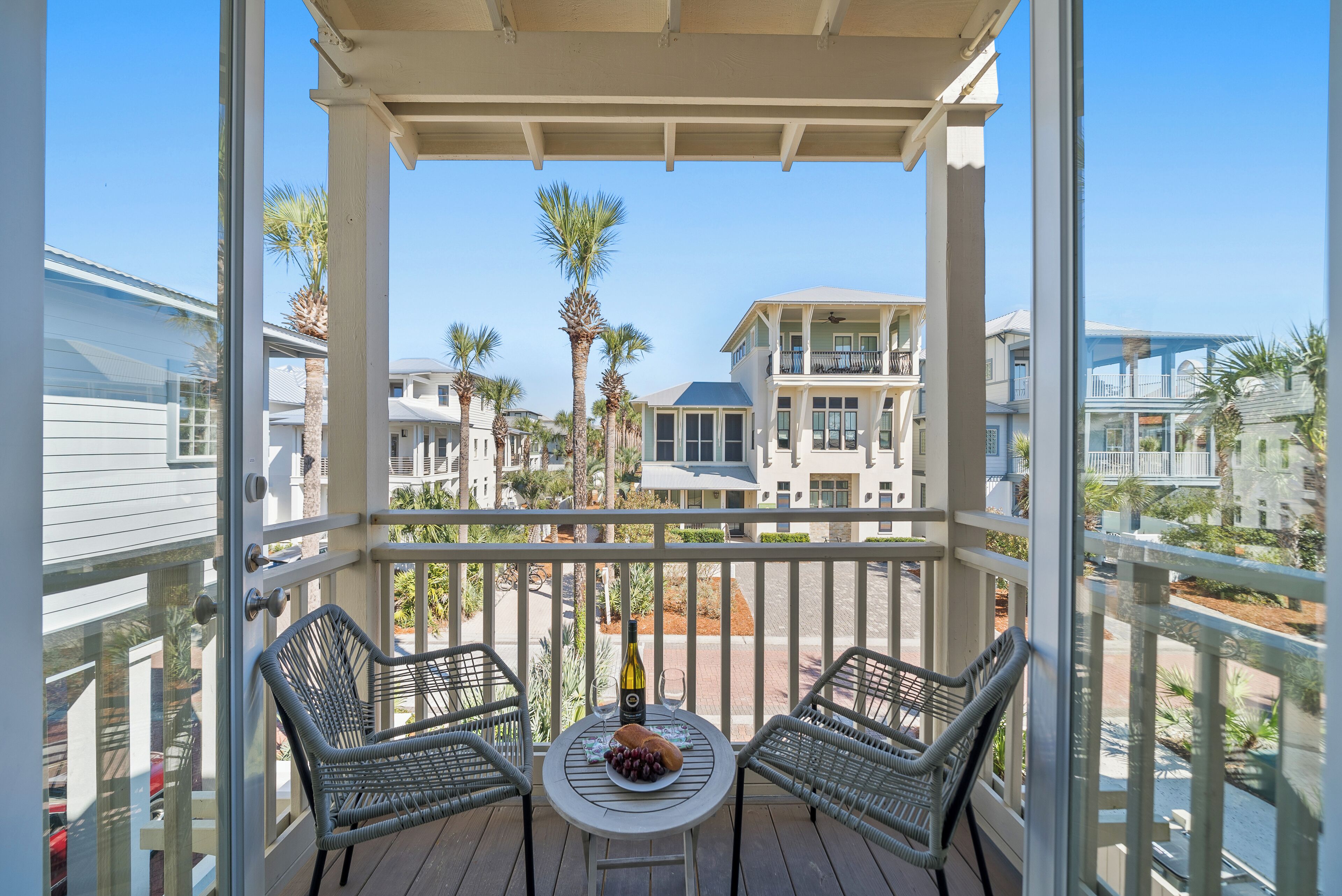 A balcony with wicker chairs, a table with a bottle and grapes, and a view of neighboring houses.