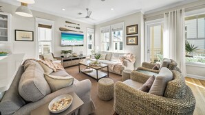 A spacious living room with a large sofa, a coffee table, and wicker chairs. There is a television and a window with curtains.