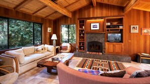 A cozy wooden interior with a fireplace, comfortable seating, and large windows offering a view of the outdoors. - Woodhaven | MCM Inspired Getaway w/ Amazing Views (Inverness)