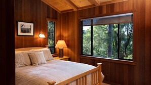 A wooden-paneled bedroom with a large bed, a nightstand with a lamp, and a window with a forest view. - Woodhaven | MCM Inspired Getaway w/ Amazing Views (Inverness)