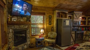 A cozy cabin interior with a stone fireplace, a television mounted on the wall, a wooden dining table, a patterned armchair, and a kitchen area with a refrigerator and microwave. - Beavers Bend State Park entrance 259A , Southern Hill Addition; Wood Knot Cabin (Broken Bow)