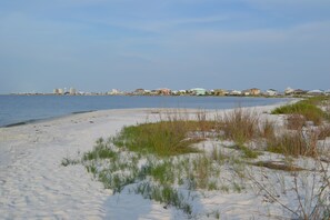 Une plage de sable avec une végétation clairsemée, une mer calme et une côte lointaine parsemée de bâtiments.